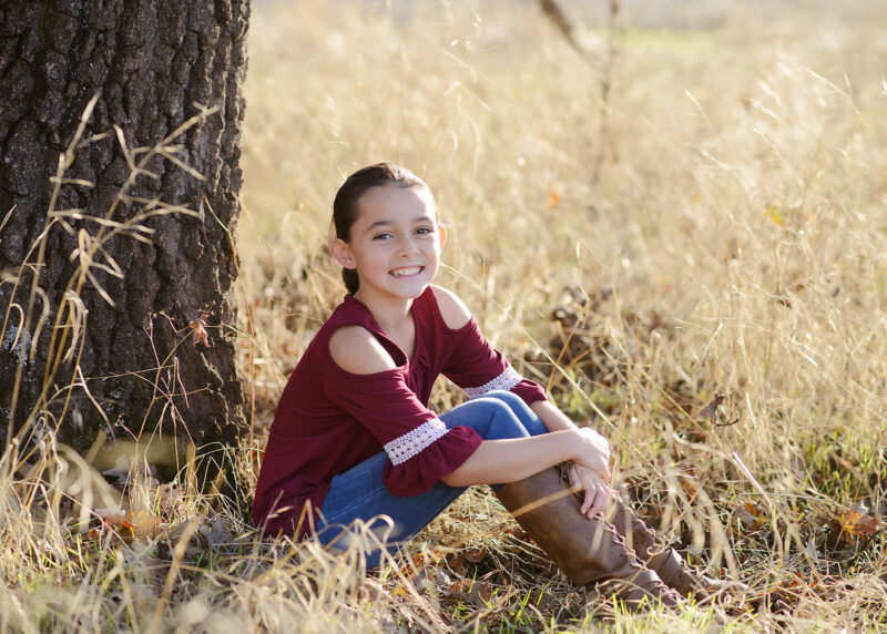 Girl wearing red top and boots sitting on dry grass under a tree in Cameron Park