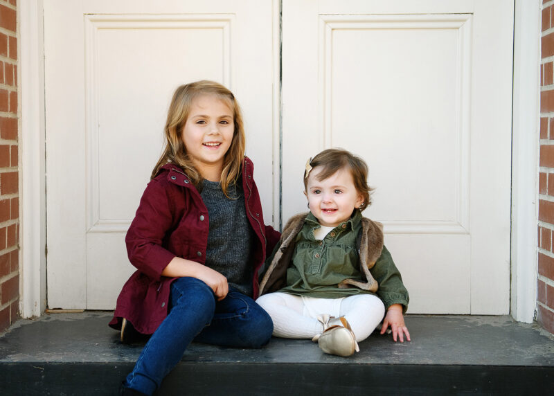 Big sister and baby sister sit on porch step against white doors in Old Sacramento