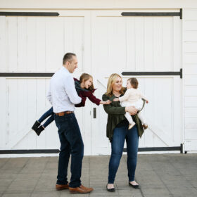 Mom and dad hold daughters as they touch hands and laugh in front of white wooden doors in Old Sacramento