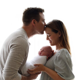 Newborn session with dad kissing mom while holding sleeping newborn baby in home