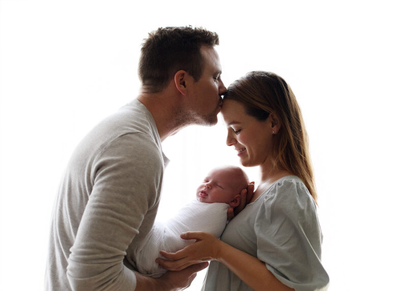 Newborn session with dad kissing mom while holding sleeping newborn baby in home