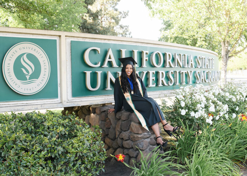 Sacramento State Senior Girl in front of university sign in cap and gown