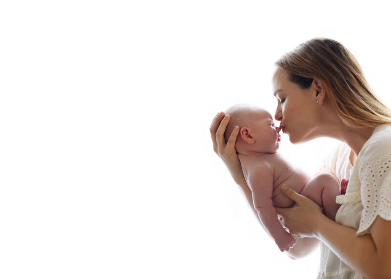 Mom kissing newborn baby boy against white light background in home