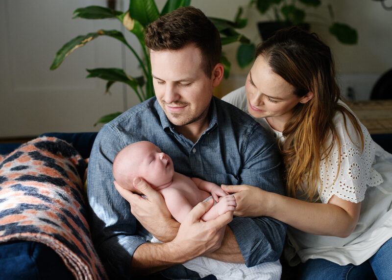 Mom and dad hold newborn baby boy while sitting on couch at home lifestyle session