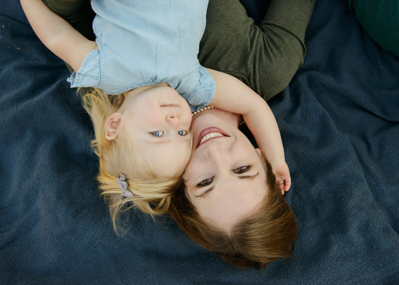 Mom and toddler daughter lie on blanket and look at camera