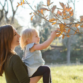 Mom holds toddler daughter as she picks a brown fall leaf on a tree in Sacramento park