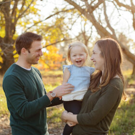Mom and dad smile while holding toddler daughter laughs with foliage in background