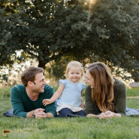 Mom and dad lie on grass while looking at toddler daughter with green trees in background Sacramento
