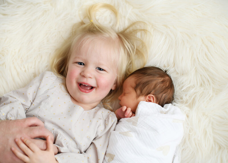 Big sister smiling and lying next to sleeping newborn sibling on sherpa throw in Sacramento studio