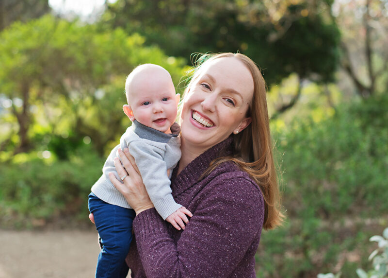 Mom smiling as she holds her baby boy against green tree background in Davis