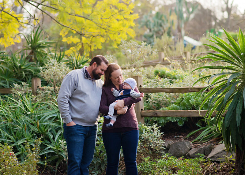Mom holds baby boy while dad looks lovingly surrounded and framed by green trees and bushes in Land Park