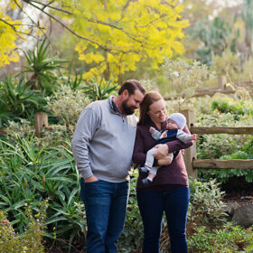 Mom holding baby son while dad smiles at him surrounded by lush greenery in Land Park