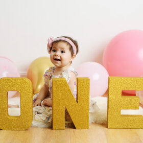 One year old baby girl smiles for camera and poses under ONE letters in Sacramento studio