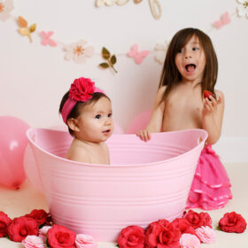 One year old girl in pink tub with strawberries and milk while big sister snacks on strawberry