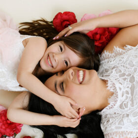 Mom and big sister cuddle close on the floor surrounded by red flowers in Sacramento studio