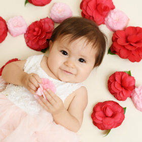 One year old girl lies down surrounded by red and pink flowers in Sacramento studio