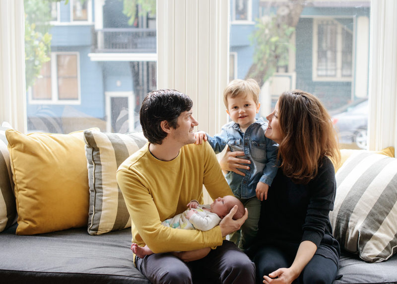 Dad holding newborn as mom holds big brother by bay windows in San Francisco