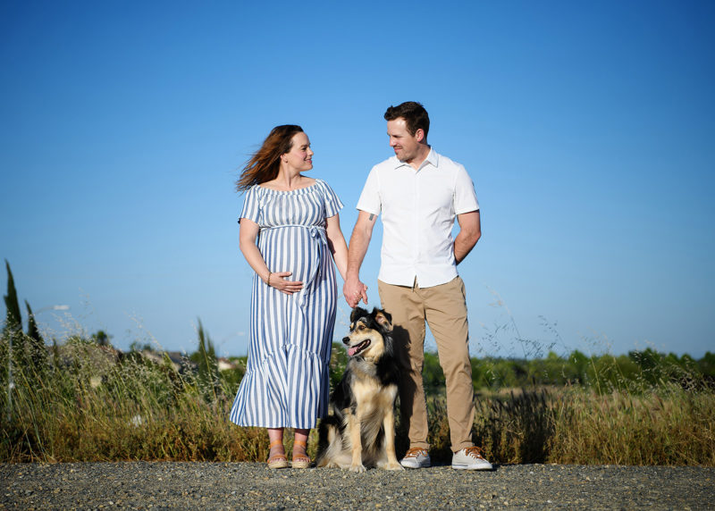 Pregnant woman and husband hold hands and look at each other lovingly with their dog in between them Sacramento