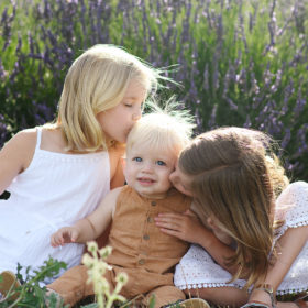 Sisters kiss baby brother on the cheek while sitting on ground in the middle of lavender field in Araceli Farms Dixon