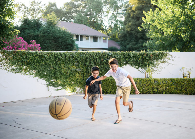 Brothers chasing after basketball in Sacramento backyard with fence growing ivy in background