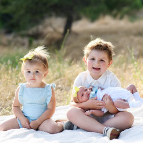 Toddler sister and big brother holding newborn baby sister on blanket on dry grass in Rocklin