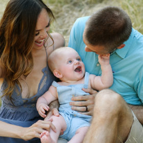 Mom and dad look at baby boy as he smiles at them while sitting on dry grass in Davis