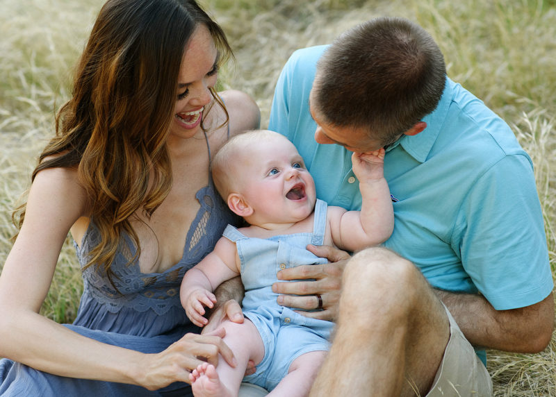 Mom and dad look at baby boy as he smiles at them while sitting on dry grass in Davis