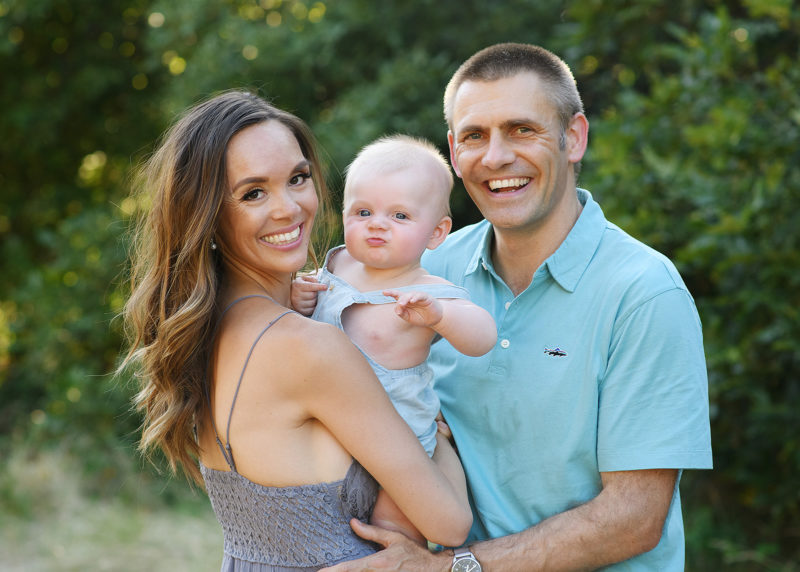 Mom and dad holding baby boy as they smile for the camera with trees in background Roseville