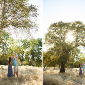Wide shot of dad and mom holding baby boy while under a large tree in dry grass field in Davis