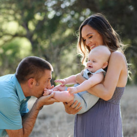 Dad kissing baby boy’s feet while mom holds him with trees and dry grass in background