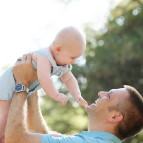 Dad lifting up baby boy in the air and smiling with trees and sky in background in Davis