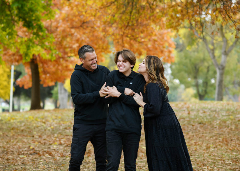 Lots of laughs as mom and dad hug son wearing all black with fall foliage background Sacramento
