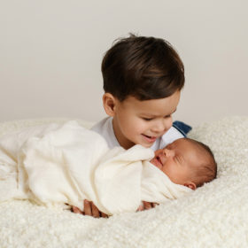 Big brother smiling at newborn baby sister in white swaddle and fuzzy blanket in Sacramento studio