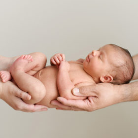 Newborn baby girl being held by mom and dad’s hands on blank wall in Sacramento studio