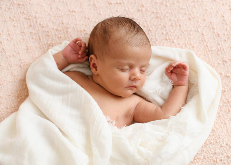 Newborn baby girl sleeping with arms by head wrapped in white muslin swaddle on pink blanket in Sacramento studio