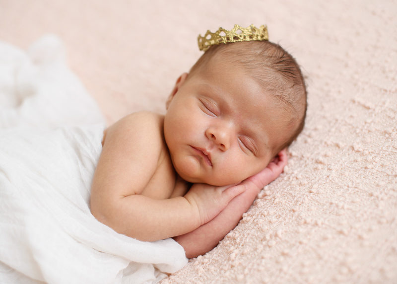 Newborn baby girl sleeping with hands under her head with tiny gold crown on head in Sacramento studio