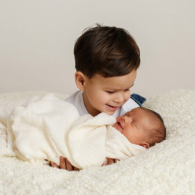 Big brother smiling at newborn baby sister in white swaddle and fuzzy blanket in Sacramento studio