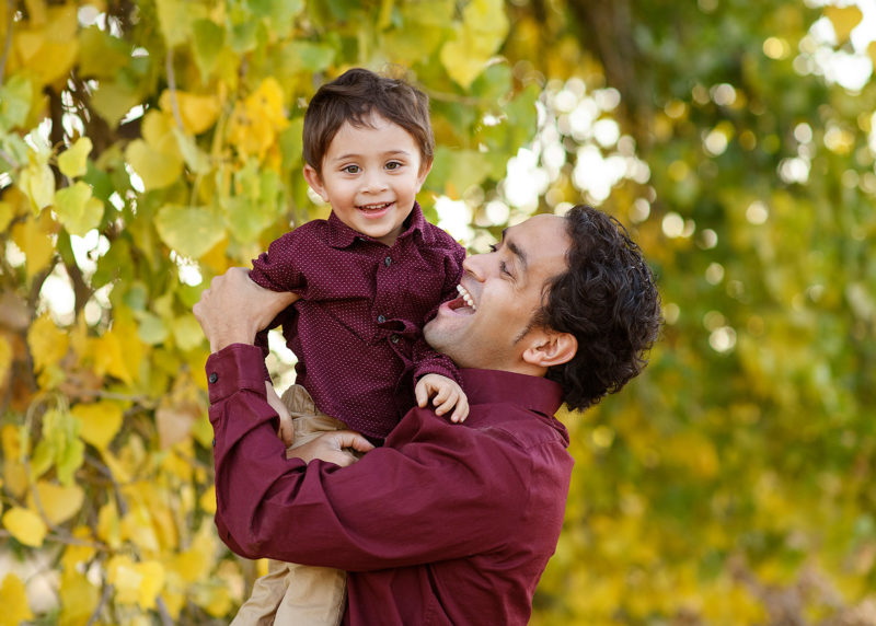 Dad smiling and holding son with yellow leaves on trees as background River Park