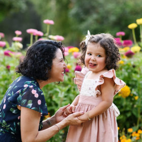 Toddler girl sticking her tongue out at the camera while mom looks at her and holds her hands by wildflowers in Sacramento Capitol