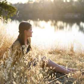 Teen girl sitting on dry grass by lake’s reflection in natural sunlight in Folsom