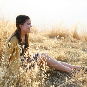 Teen girl sitting and smiling on dry grass by lake’s reflection in natural sunlight in Folsom