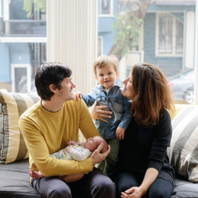 Dad holding newborn as mom holds big brother by bay windows in San Francisco
