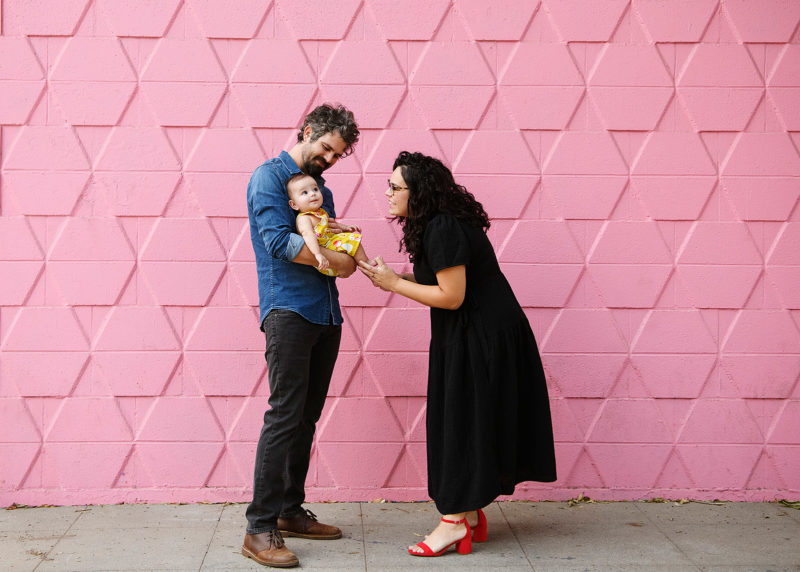 Mom looks at baby daughter as dad holds her against a pink tiled wall in midtown Sacramento