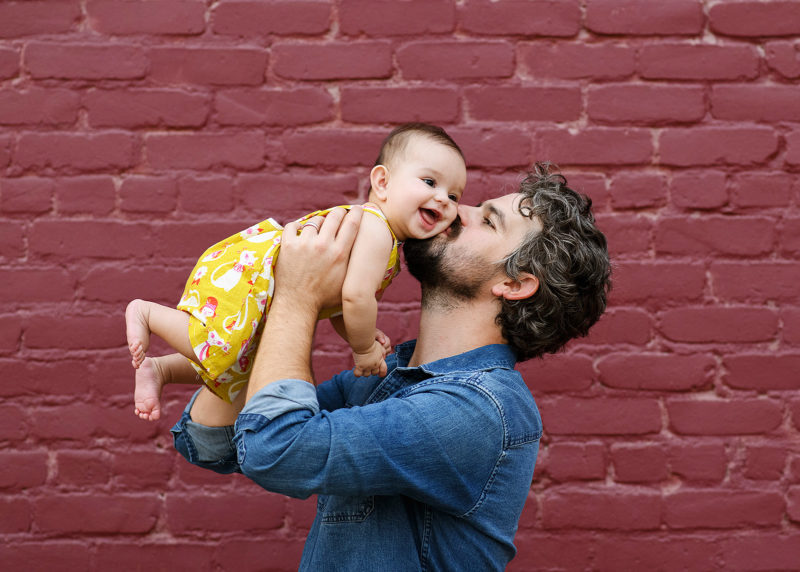Dad kissing baby daughter on cheek as she smiles against purple brick background in Sacramento