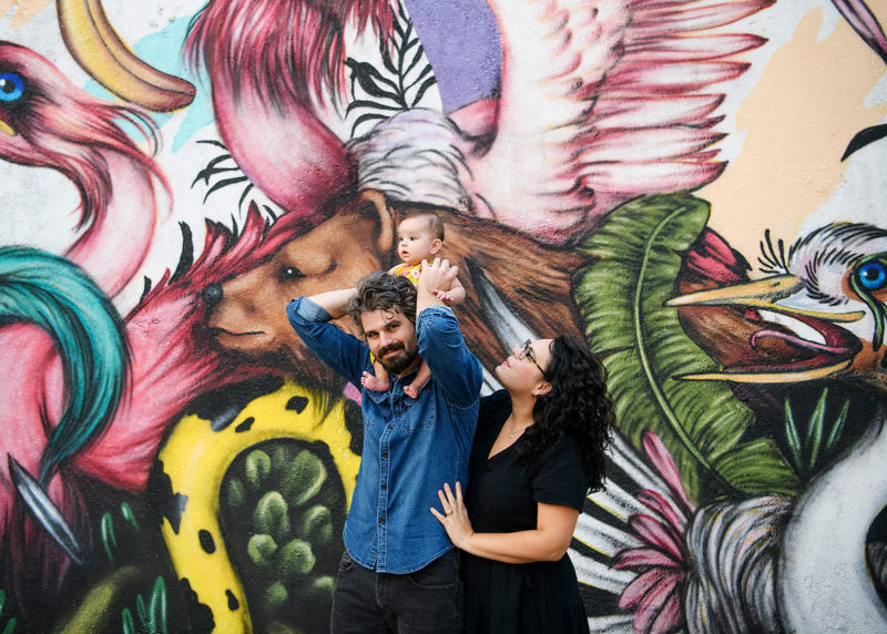 Dad carrying baby daughter on shoulders as mom watches in front of Sacramento mural