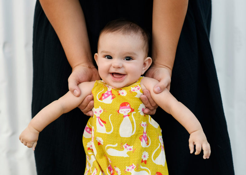 Baby girl smiling directly at camera wearing yellow romper as mom supports her in Sacramento