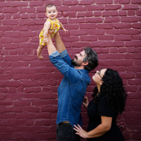 Dad lifting up baby girl while mom watches with purple brick background in Sacramento