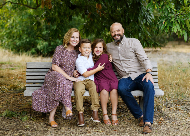 Family sitting on bench and smiling beneath a large green tree in Sacramento