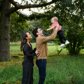 Dad holding up son in the air as mom smiles surrounded by large tree and green grass in Sacramento
