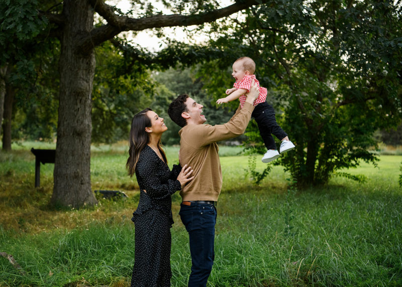 Dad holding up son in the air as mom smiles surrounded by large tree and green grass in Davis